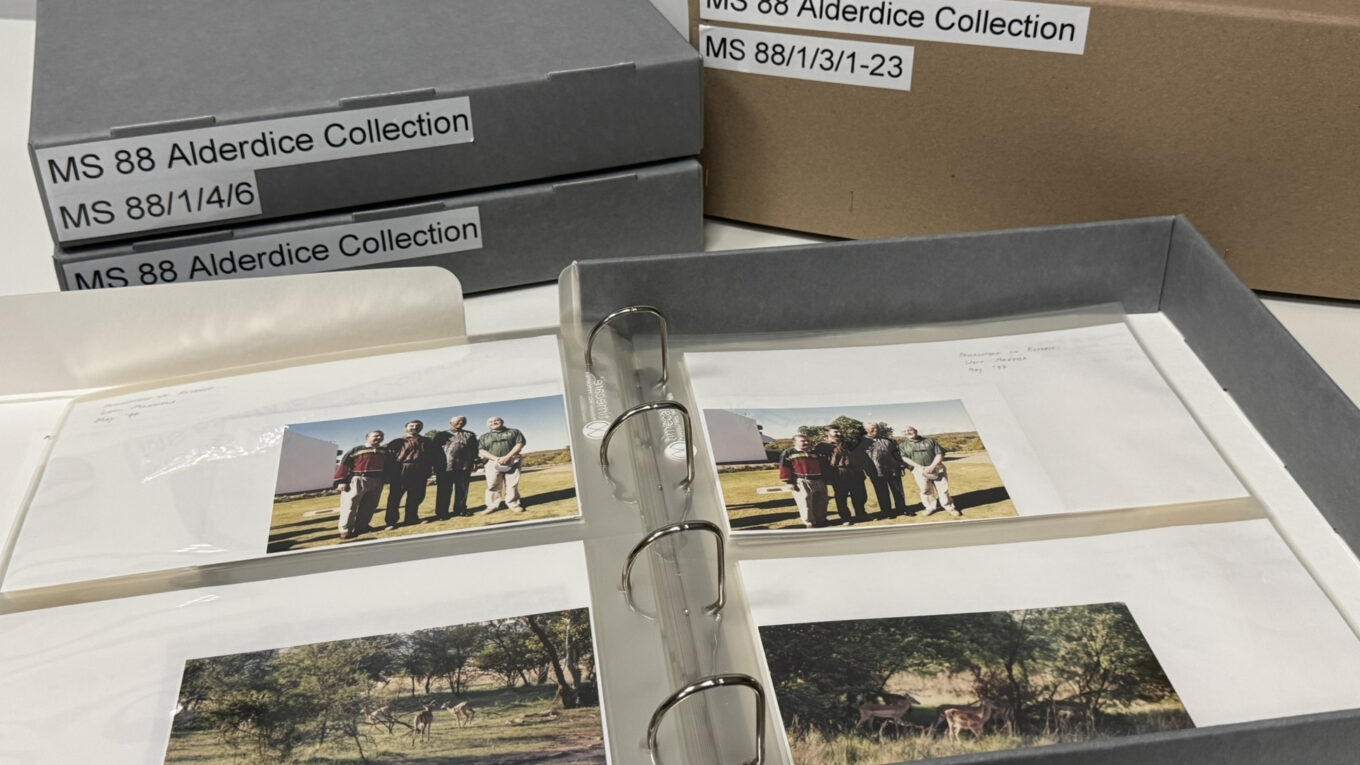 Photograph of archival boxes and photo albums labelled "MS 88 Alderdice Collection". An open archival album shows photographs of Lord Alderdice's visit to South Africa in May 1997, including images of wildlife and a meeting with Nelson Mandela.